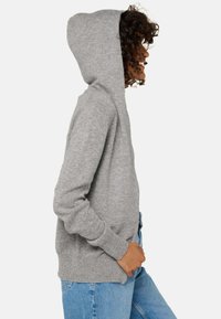 Sweat à capuche gris fabriqué en tissu doux et texturé. Il comporte une poche frontale et des poignets côtelés, porté par-dessus un jean bleu clair.