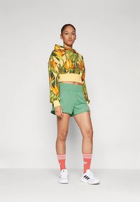 Sudadera con capucha recortada con diseño floral amarillo, combinada con pantalones cortos verdes, calcetas a rayas rosas y zapatillas blancas. Tela suave, ajuste casual.