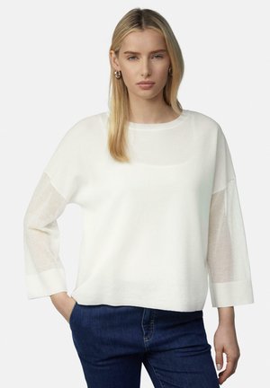 White oversized sweater with sheer long sleeves and a round neck, paired with blue denim jeans. The fabric appears soft and lightweight.