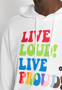 Vit hoodie med färgglada, djärva bokstäver "LEVA HÖGT! LEVA STOLT" buren av en person med mörk hud, som visar överkroppen och nacken endast.