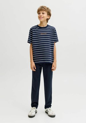 Boy wearing a navy striped T-shirt, dark pants, and white sneakers, standing against a plain light background, smiling and looking right.