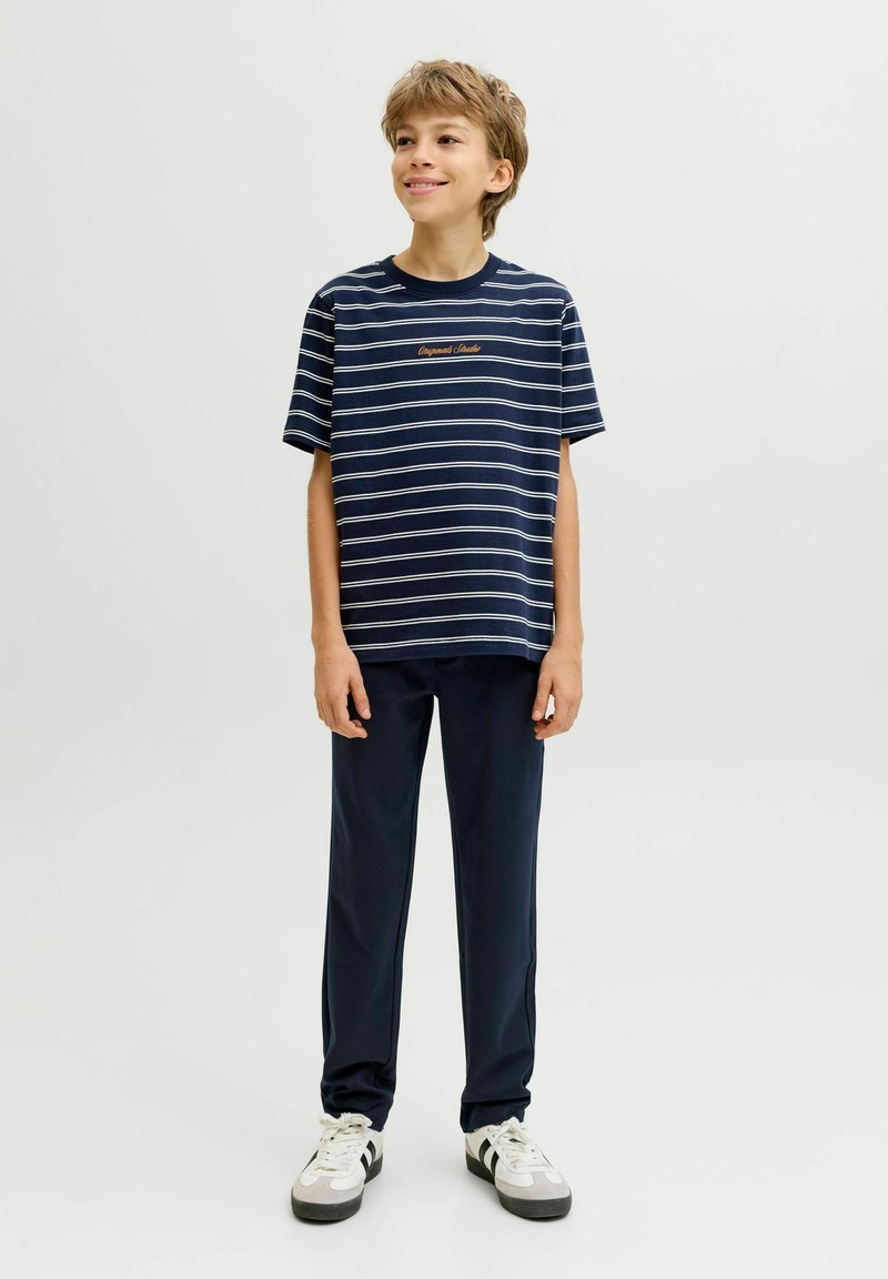 Garçon portant un t-shirt rayé marine, un pantalon foncé et des baskets blanches, debout devant un fond clair uni, souriant et regardant vers la droite.