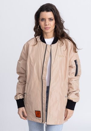 Licht beige bomberjack met een ritssluiting aan de voorkant, ribbelige zwarte manchetten en een oranje label op de linkse zoom; gemaakt van winddicht, glanzend materiaal.