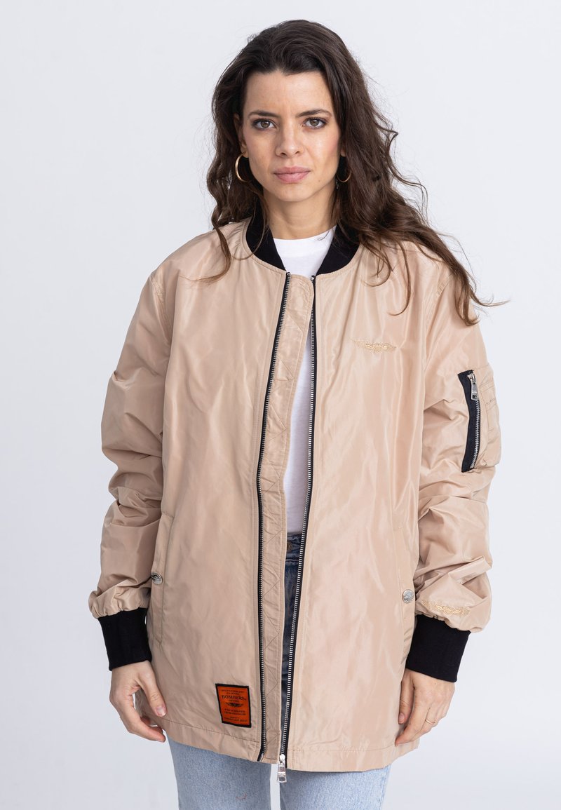 Veste bomber beige clair avec une fermeture éclair à l'avant, des poignets noirs côtelés et une étiquette orange sur l'ourlet gauche ; fabriquée en tissu imperméable au vent et brillant.