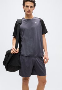 Νέος άνδρας που φορά γκρι και μαύρο αθλητικό μπλουζάκι και σορτς Puma, κρατώντας μια μαύρη τσάντα γυμναστηρίου στον ώμο του, με απλό φόντο.