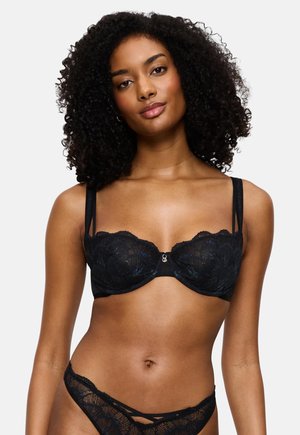 Mulher com cabelo encaracolado a fazer de modelo com um sutiã de renda preto e cuecas de renda a condizer, em fundo branco liso.