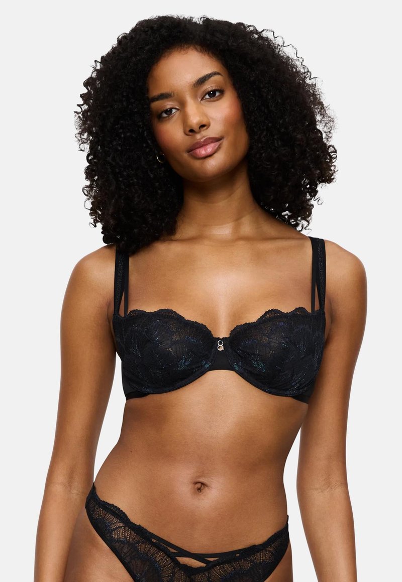 Γυναίκα με σγουρά μαλλιά ποζάρει φορώντας μαύρο δαντελένιο σουτιέν και συνδυασμένα δαντελένια εσώρουχα πάνω σε λευκό φόντο.