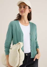 Ljusgrön cardigan med knappar fram, ribbade ärmändar och fickor, kombinerad med en vit t-shirt och håller en krämfärgad väska.
