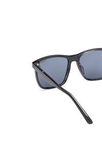 Mustat suorakulmion muotoiset aurinkolasit, joissa on polarisoidut siniset linssit. Paksut kehykset ja hoikat varret, joissa on hillitty brändäys sivuilla. Sileä pinta.