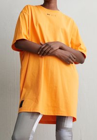 Oranžové oversized tričko vyrobené z bavlny, se zaobleným výstřihem a krátkými rukávy. Malé černé logo je umístěno poblíž límce.