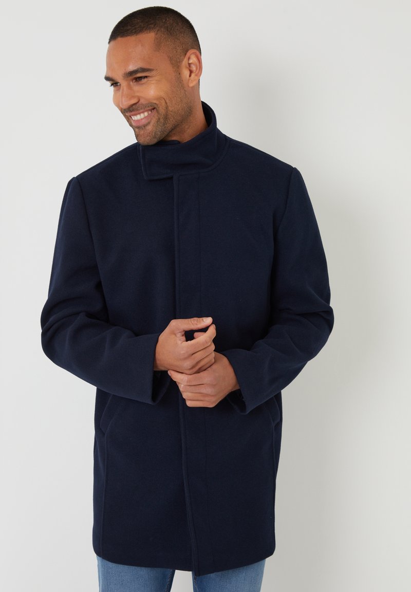 Marineblauer Wollmantel mit hohem Kragen, langen Ärmeln und geradem Schnitt, ausgestattet mit seitlichen Taschen und einem sauberen, minimalistischen Design.