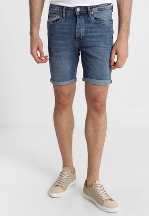 Mężczyzna w niebieskich dżinsowych szortach z podwiniętymi nogawkami i beżowych sneakersach, stojący na tle białej ściany.