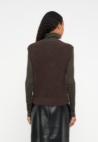 Kobieta z długimi, kręconymi włosami, ubrana w brązowy bezrękawnik z pluszu na oliwkowym golfie i czarnej skórzanej spódnicy, zwrócona plecami.