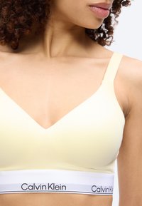 Κοντινό πλάνο γυναίκας που φοράει ανοιχτό κίτρινο αθλητικό σουτιέν Calvin Klein με ρυθμιζόμενες τιράντες και λάστιχο με το λογότυπο κάτω από το στήθος.