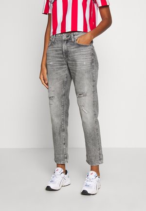 Persoon draagt grijze versleten spijkerbroek, witte sneakers en een rood-wit gestreept cropped shirt, staand tegen een effen achtergrond.