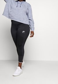 Hellgraue, kurze Kapuzenpullover mit einer frontalen Tasche, kombiniert mit schwarzen Leggings mit Textur und einem weißen Nike-Logo. Getragen mit weißen Sneakers.