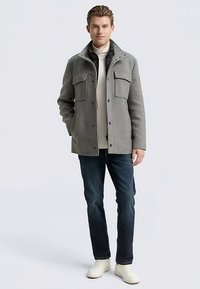 Grijze jas van een wolmix met een kraag, knopen aan de voorkant en twee grote borstzakken. Gecombineerd met een lichte trui, spijkerbroek en witte schoenen.