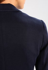 Detailní záběr na horní část zad osoby v tmavě námořnickém texturovaném blejzru na světlém jednofonním pozadí.