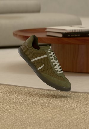 Olivengrøn sneaker med mesh- og ruskindsmaterialer, med hvide accenter, rund tå og tekstureret gummisål, vist svævende over et tæppe.