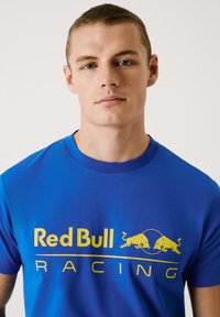 Kuning sinine puuvillane T-särk, millel on suur kollane "Red Bull Racing" logo ja kaks kollast pulli graafikat, ümmargune kaelus ja lühikesed varrukad.
