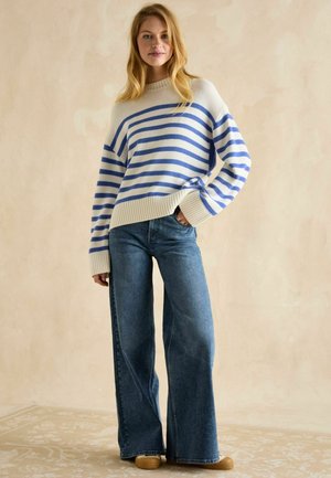Striped sweater in cream and blue, oversized fit, ribbed cuffs and hem. Paired with wide-leg blue jeans and tan slip-on shoes.