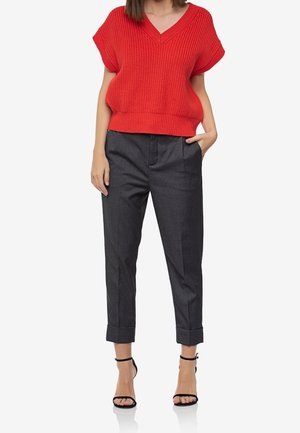 Kvinde iført en rød strikket ærmeløs top med V-hals, mørkegrå bukser med opsmøg, og sorte høje hæle med ankelsrem.