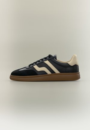 Basket noire avec des rayures beige et un talon, semelle marron texturée et lacets noirs sur un fond neutre.