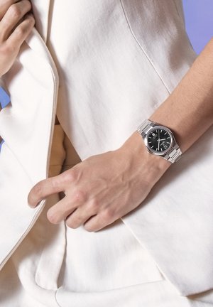 Orologio da polso argentato con quadrante nero, cinturino in acciaio inossidabile e display della data, indossato su una mano che tiene un capo bianco. Tessuto texturizzato visibile.