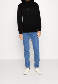 Černá bavlněná mikina s kapucí na šňůrku a přední kapsou, v kombinaci se světle modrými džíny a bílými teniskami. Jednoduchý design, volný střih.