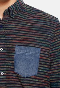 Randig skjorta med flerfärgade vertikala linjer; har en denimfickor på vänster sida och en ståkrage.