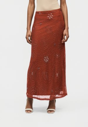 Φούστα μακριά σε χρώμα σκουριάς με λουλουδάτα σχέδια από χάντρες και παγιέτες, φορεμένη με λευκές πέδιλες ανοιχτές στα δάχτυλα και μπεζ αμάνικη μπλούζα.