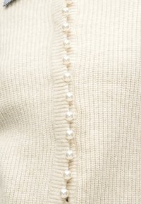 Cardigã creme em malha com botões redondos de pérola ao longo do centro. O tecido apresenta uma aparência texturada. Detalhe de colarinho cinzento claro visível.