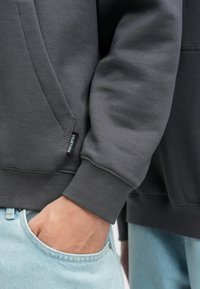 Sötétszürke pulóver puha tapintású anyagból, kenguruzsebbel és bordázott mandzsettákkal. Tartalmaz egy kis fekete címkét a "YOURTURN" felirattal.