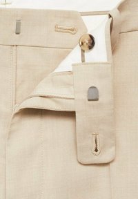 Light beige trousers with a smooth texture, featuring a concealed zipper, a brown button, and a fabric pocket for secure closure.