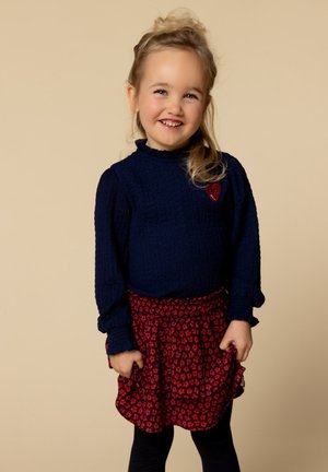 Lachend jong meisje met blond haar draagt een marineblauwe longsleeve top en een rode bloemenrok, terwijl ze tegen een beige achtergrond staat.