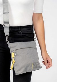 Borsa a tracolla grigia realizzata in tessuto testurizzato. Caratterizzata da una tasca frontale con zip, un accento giallo sulla cerniera e una tracolla grigia regolabile.