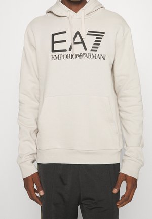 Mann trägt einen cremefarbenen Hoodie mit schwarzem "EA7 Emporio Armani"-Logo, kombiniert mit dunkler Hose, steht vor schlichtem Hintergrund.