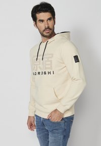 Sudadera color crema con un gran estampado de texto negro, capucha ajustable con cordones, bolsillo tipo canguro en el frente y un pequeño parche de logo negro en la manga.