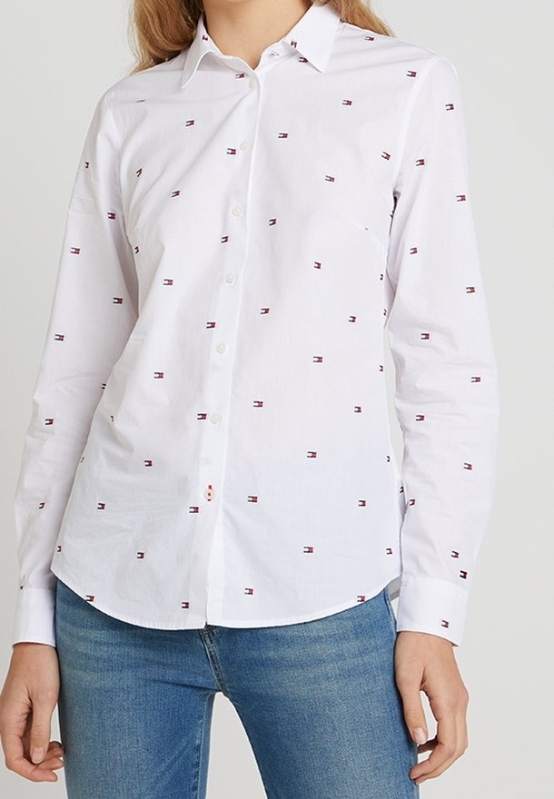 Camisa blanca de botones con pequeños patrones de emblemas rojos y azules, diseño con cuello y mangas largas, combinada con jeans azules.