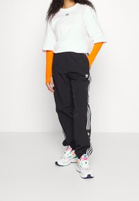 Persona con una maglietta bianca Adidas sopra maniche lunghe arancioni, pantaloni da jogging neri Adidas e scarpe multicolori bianche, in piedi con una mano in tasca.