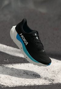 Schwarzer und blauer Hoka-Laufschuh mit dicker weißer Sohle, der in der Luft über einer Asphaltoberfläche abgebildet ist.