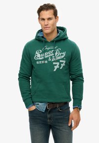 Haut à capuche vert avec cordons de serrage, poche avant et impression de logo blanc. Porté par-dessus une chemise en denim, avec un jean bleu et une ceinture marron.