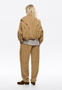 Una persona con capelli mossi sta in piedi di spalle rispetto alla fotocamera, indossando un'ampia giacca beige, un maglione grigio e sabot marroni.