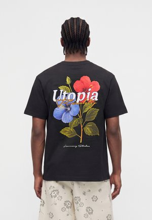 Man met gevlochten haar die een zwart T-shirt draagt met rode en blauwe bloemen en de tekst "Utopia Reality" op de rug, gecombineerd met een korte broek met bloemenprint.