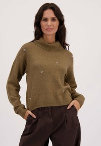 Olijfgroene trui met hoge kraag, voorzien van een zachte textuur, brede mouwen en kleine zilveren versieringen door de stof.