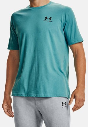 Mann trägt ein türkisfarbenes Under Armour Kurzarmshirt und hellgraue Hose mit Under Armour-Logos auf Brust und Oberschenkel, steht mit entspannten Händen.