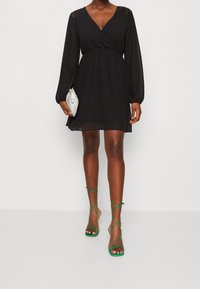 Robe portefeuille noire à manches longues avec détail en dentelle aux épaules. Associée à des sandales vertes à lanières et une pochette argentée.