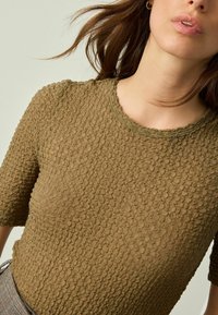 Olivgrünes, strukturiertes Strickoberteil mit kurzen Ärmeln, rundem Ausschnitt und einem dezenten Muster, das eine weiche, leicht erhabene Oberfläche zeigt.