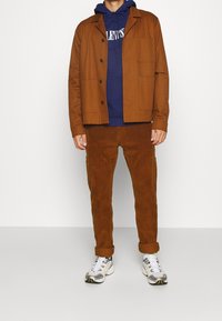 Giacca in corduroy marrone su una felpa blu navy, abbinata a pantaloni in corduroy marrone e sneakers bianche con accenti metallici.