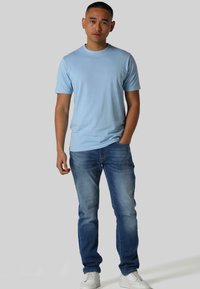 Svijetloplava pamučna majica s izvezenim tekstom, u kombinaciji sa svijetloplavim trapericama i bijelim tenisicama. Model stoji s rukama u džepovima.